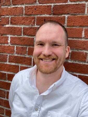 Picture of Ryan McCarthy, a man with short dark hair and a short reddish beard. He is smiling. He is wearing a white oxford shirt and leaning against a brick wall.